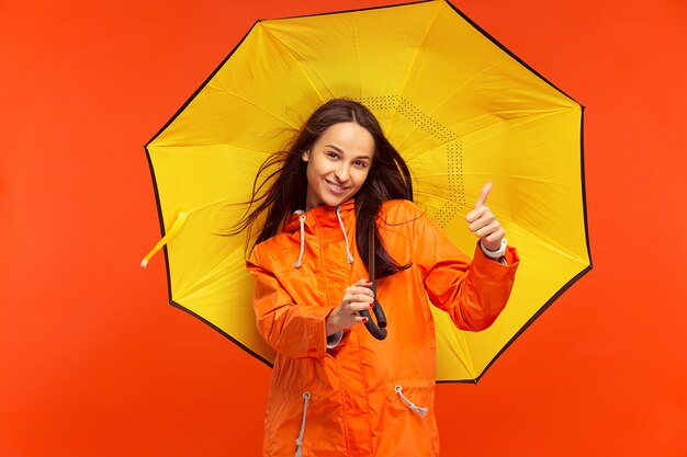 Het gelukkige glimlachende jonge meisje stellen bij studio in de herfst oranje jasje dat op rood wordt geïsoleerd. Menselijke positieve emoties. Concept van het koude weer. Vrouwelijke mode-concepten