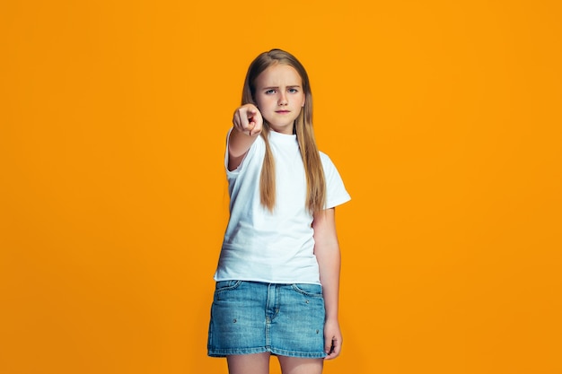 Het gelukkige tienermeisje wijst naar u, het portret van de halve lengteclose-up op oranje achtergrond.