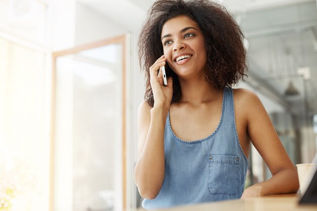 Het gelukkige vrolijke mooie Afrikaanse vrouw glimlachen die op telefoonzitting spreken in koffie.