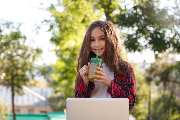 Het glimlachen jonge donkerbruine vrouwenzitting in park met laptop computer