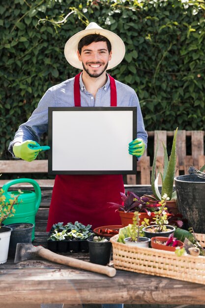 Het glimlachen portret van een jonge mens die zich achter de ingemaakte installaties bevinden die de vinger op wit leeg frame richten