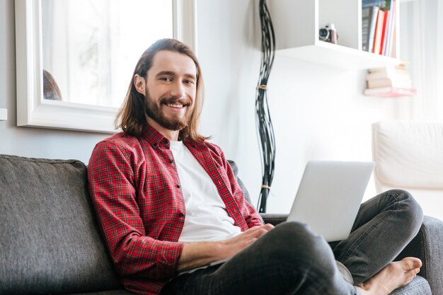 Het glimlachen van gebaarde jonge mensenzitting op laag en het gebruiken van laptop