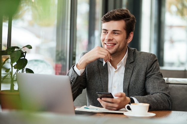 Het glimlachen zakenmanzitting door de lijst in koffie met laptop computer en smartphone terwijl weg het kijken
