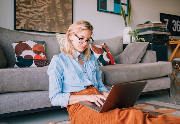 Het glimlachen zitting van de blonde jonge vrouw dichtbij de bank die laptop met behulp van