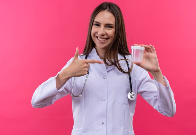 Het glimlachende jonge artsenmeisje die stethoscoop medische toga dragen wijst naar leeg blik in haar hand op geïsoleerde roze achtergrond