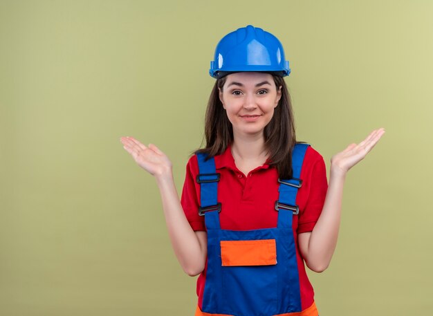Het glimlachende jonge bouwersmeisje met blauwe veiligheidshelm houdt hand op geïsoleerde groene achtergrond met exemplaarruimte vast