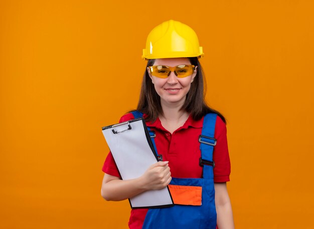 Het glimlachende jonge bouwersmeisje met veiligheidsbril houdt klembord op geïsoleerde oranje achtergrond