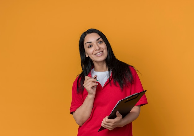 Het glimlachende jonge donkerbruine kaukasische meisje dat rood overhemd draagt houdt pen en klembord dat op oranje muur wordt geïsoleerd