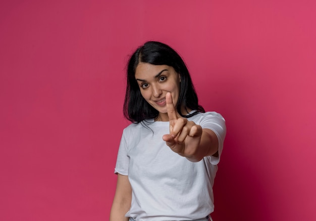 Het glimlachende jonge donkerbruine Kaukasische meisje toont wijsvinger die op roze muur wordt geïsoleerd