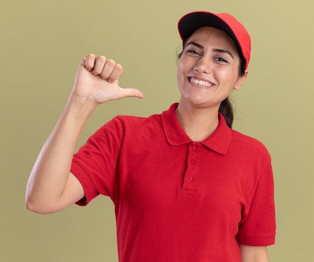 Het glimlachende jonge leveringsmeisje die eenvormig met GLB dragen wijst naar zichzelf geïsoleerd op olijfgroene muur