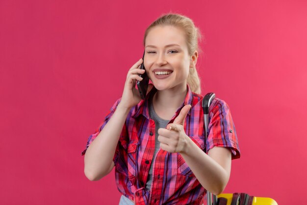 Het glimlachende jonge meisje dat van de reiziger een rood overhemd draagt, spreekt via de telefoon toont u gebaar op geïsoleerde roze achtergrond