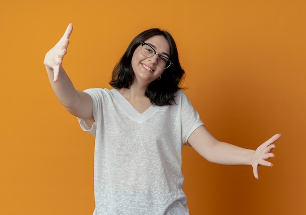 Het glimlachende jonge vrij Kaukasische meisje dat glazen draagt beweert iets te houden dat op oranje achtergrond wordt geïsoleerd