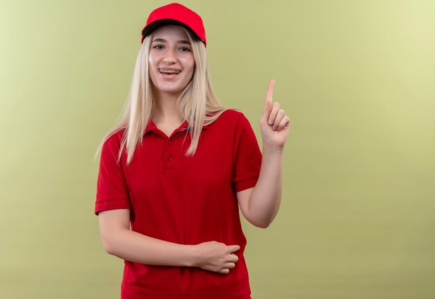 Het glimlachende levering jonge meisje dat rode t-shirt en pet in tandsteun draagt wijst omhoog op geïsoleerde groene achtergrond