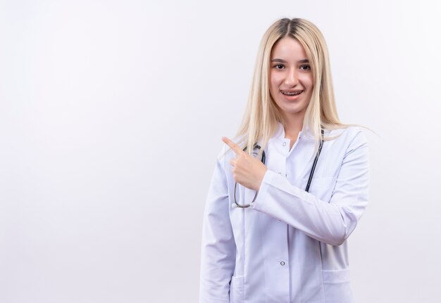 Het glimlachende meisje dat van het artsen jonge blonde stethoscoop en medische toga in tandsteun draagt wijst naar kant op geïsoleerde witte achtergrond