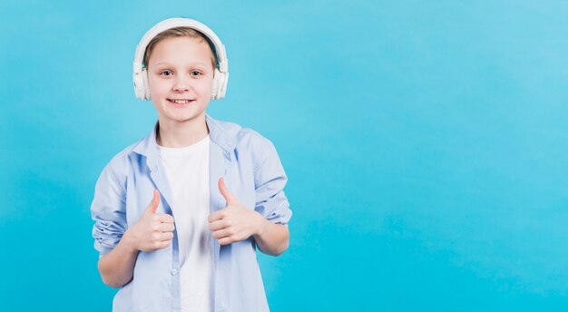 Het glimlachende portret van een jongen met witte hoofdtelefoon op zijn hoofd die duim tonen ondertekent omhoog tegen blauwe achtergrond