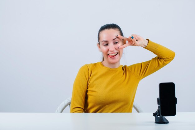 Het grappige bloggermeisje stelt door twee gebaar te tonen en tong uit te steken voor mobiele camera op witte achtergrond