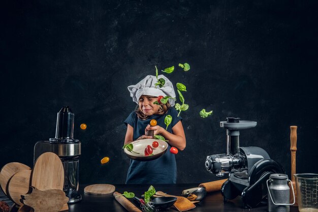 Het grappige meisje gooit groenten op de pan bij donkere fotostudio.