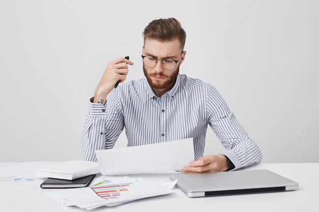 Het horizontale portret van aantrekkelijke geslagen mannelijke manager, zit op kantoor, omringd met laptop