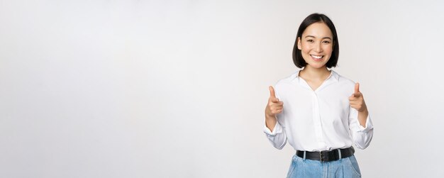 Het is je gefeliciteerd Glimlachende aantrekkelijke Aziatische vrouw zakenvrouw wijzende vingers op camera met blij gezicht complimenteren uitnodigend u permanent over witte achtergrond