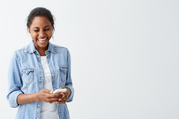 Het jonge Afro-Amerikaanse vrouw lachen, die gelukkige uitdrukking heeft terwijl het lezen van berichten van haar vrienden die over witte muur worden geïsoleerd. Mensen en technologie concept