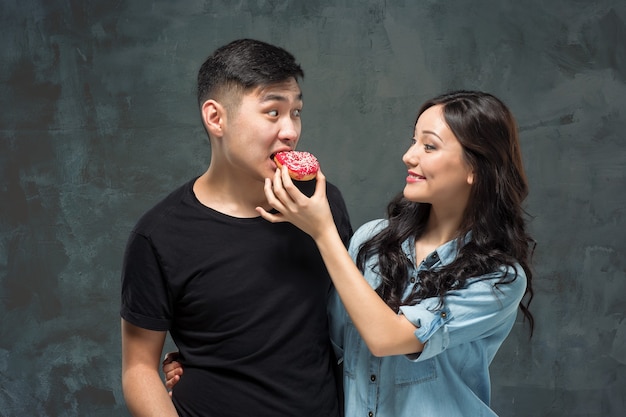 Het jonge Aziatische paar geniet van het eten van zoete kleurrijke doughnut op grijze studioachtergrond