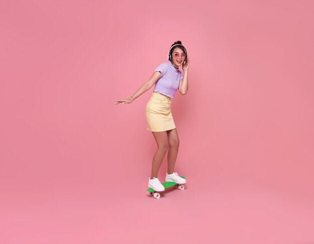 Het jonge Aziatische tienermeisje spelen op skateboard met het dragen van draadloze hoofdtelefoons die aan muziek op roze muur luisteren.