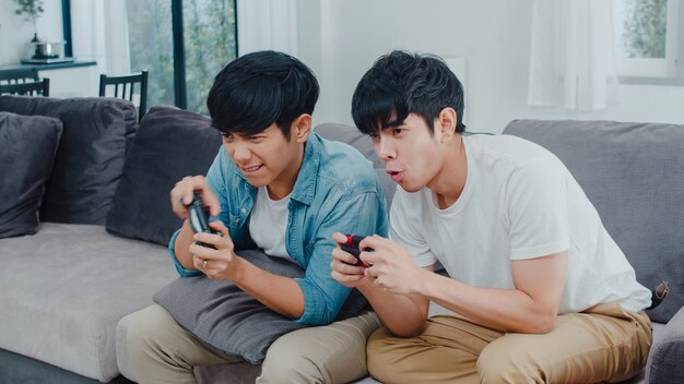 Het jonge Aziatische vrolijke paar speelt thuis spelen, Tien Koreaanse LGBTQ-mannen die joystick gebruiken die grappig gelukkig ogenblik samen op bank in woonkamer hebben bij huis.