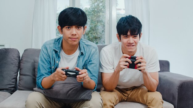 Het jonge Aziatische vrolijke paar speelt thuis spelen, Tien Koreaanse LGBTQ-mannen die joystick gebruiken die grappig gelukkig ogenblik samen op bank in woonkamer hebben bij huis.