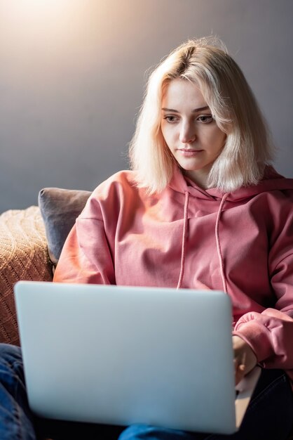 Het jonge blonde meisje van de maker van inhoud zit op haar laptop op de bank. Werken vanuit huis