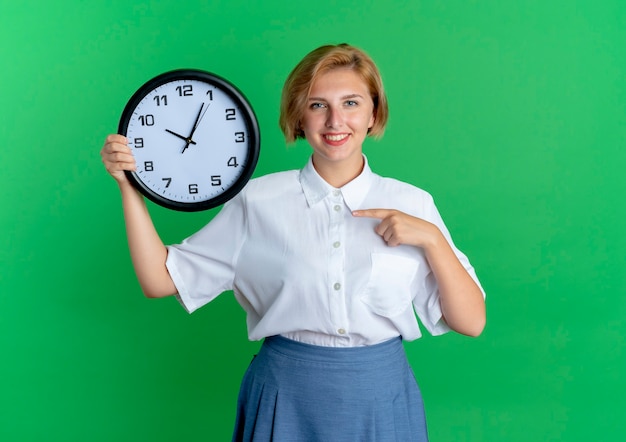 Het jonge glimlachende blonde russische meisje houdt en wijst op klok die op groene achtergrond met exemplaarruimte wordt geïsoleerd