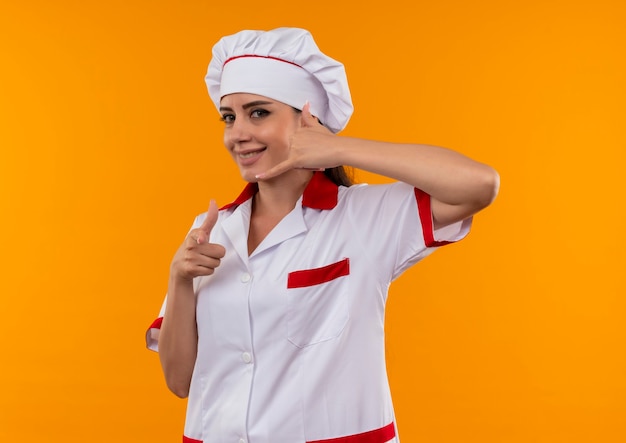 Het jonge glimlachende Kaukasisch kokmeisje in chef-kok uniforme gebaren noemt me handteken dat op oranje muur met exemplaarruimte wordt geïsoleerd