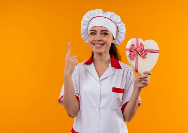 Het jonge glimlachende Kaukasische kokmeisje in eenvormige chef-kok houdt de doos van de hartvorm en wijst omhoog geïsoleerd op oranje muur met exemplaarruimte