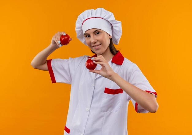 Het jonge glimlachende Kaukasische kokmeisje in eenvormige chef-kok houdt tomaten die op oranje achtergrond met exemplaarruimte worden geïsoleerd