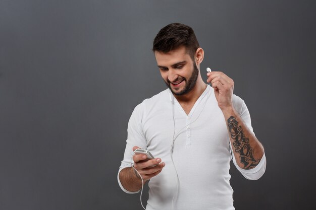 Het jonge knappe mens glimlachen die telefoon over grijze muur bekijken