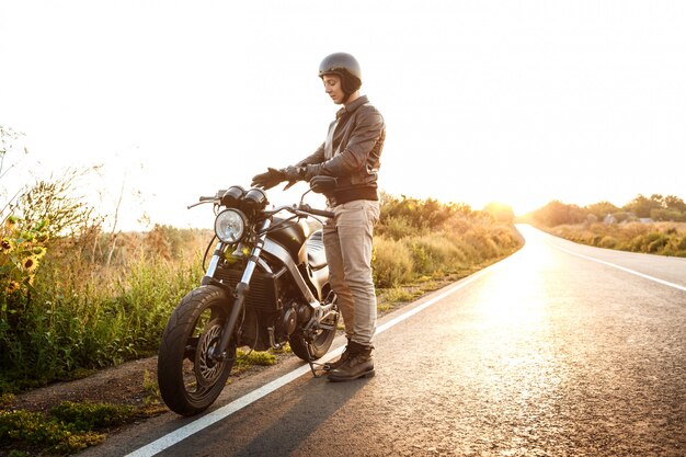 Het jonge knappe mens stellen dichtbij zijn motor bij plattelandsweg.