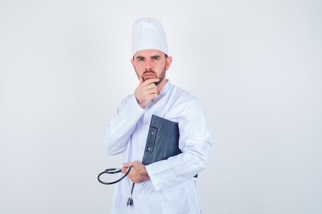 Het jonge mannelijke klembord, de stethoscoop van de artsenholding, die hand op kin in wit uniform houdt en nadenkend kijkt. vooraanzicht.