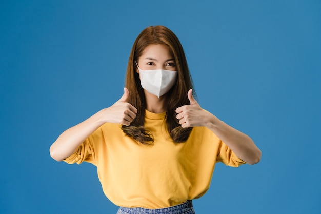 Het jonge meisje dat van Azië medisch gezichtsmasker draagt dat duim toont met gekleed in casual doek en kijkt naar camera geïsoleerd op blauwe achtergrond. Zelfisolatie, sociale afstand nemen, quarantaine voor coronavirus.