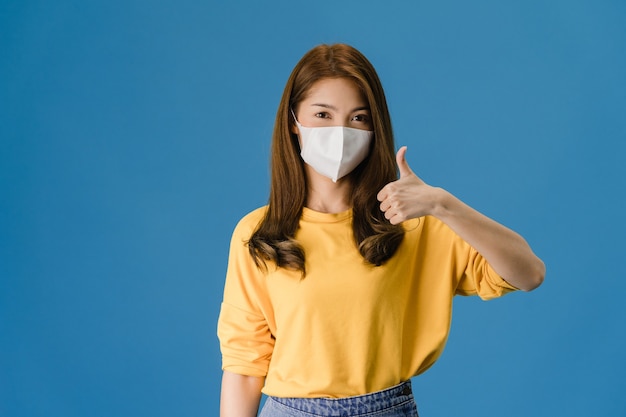 Het jonge meisje dat van azië medisch gezichtsmasker draagt dat duim toont met gekleed in casual doek en kijkt naar camera geïsoleerd op blauwe achtergrond. zelfisolatie, sociale afstand nemen, quarantaine voor coronavirus.