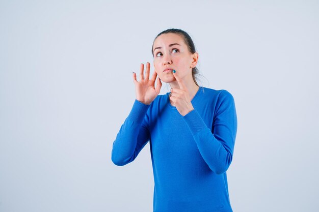 Het jonge meisje denkt door de wijsvinger op de lippen te houden en de andere hand achter het oor te plaatsen op een witte achtergrond