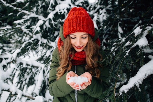 Het jonge meisje in rode gebreide de winterhoed en sjaal houdt sneeuw binnen de winter indient
