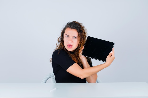 Het jonge meisje kijkt naar de camera door het tabletscherm op een witte achtergrond te tonen