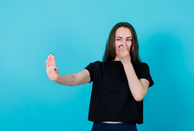 Het jonge meisje toont een stopgebaar en houdt de neus vast met de andere hand op een blauwe achtergrond