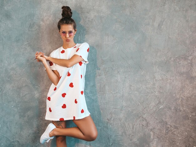 Het jonge mooie glimlachende vrouw kijken. Trendy meisje in casual witte zomerjurk en zonnebril.