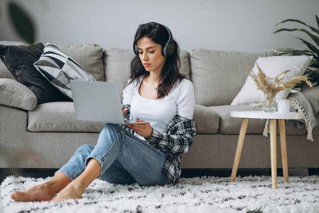 Het jonge mooie huis die van de vrouwenzitting aan laptop in oortelefoons werken