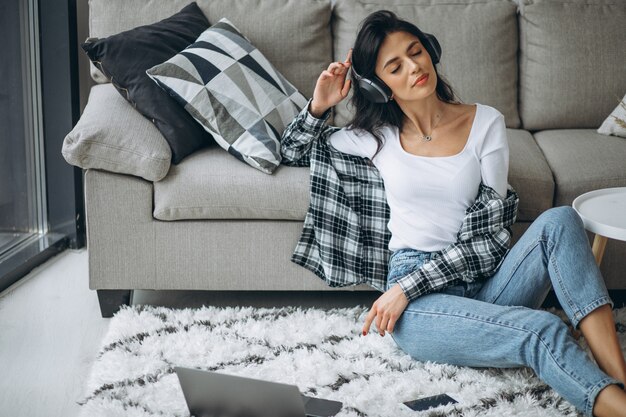 Het jonge mooie huis die van de vrouwenzitting aan laptop in oortelefoons werken