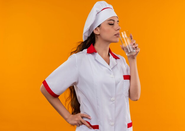 Het jonge tevreden Kaukasisch kokmeisje in eenvormige chef-kok beweert glas water te drinken dat op oranje muur met exemplaarruimte wordt geïsoleerd