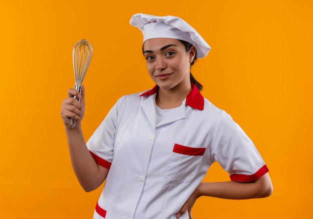 Het jonge tevreden kaukasische kokmeisje in eenvormige chef-kok houdt klopper en legt hand op taille die op oranje muur met exemplaarruimte wordt geïsoleerd