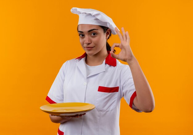 Het jonge tevreden Kaukasische kokmeisje in eenvormige chef-kok houdt plaat en gebaren ok handteken dat op oranje muur met exemplaarruimte wordt geïsoleerd