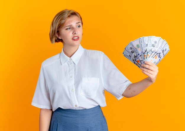 Het jonge verwarde blonde Russische meisje houdt en bekijkt geld dat op oranje achtergrond met exemplaarruimte wordt geïsoleerd