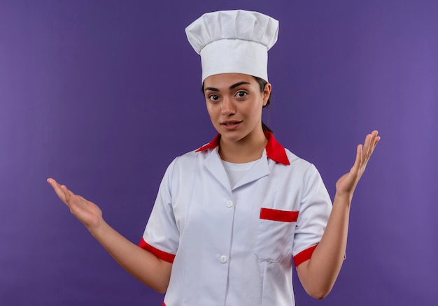 Het jonge verwarde kaukasisch kokmeisje in eenvormige chef-kok houdt handen omhoog geïsoleerd op violette muur met exemplaarruimte